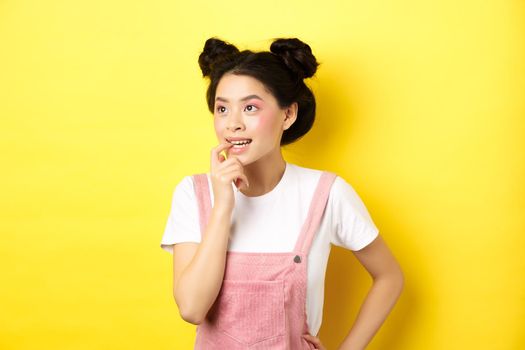Pensive asian girl feeling excited, biting fingernail and looking left thoughtful, making choice, standing in summer clothes against pink background.