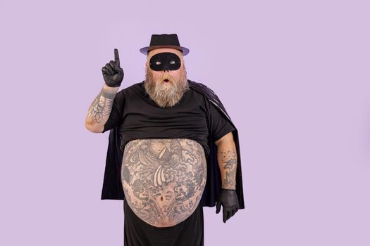 Surprised mature man with overweight in black hero suit with cape and mask got idea and points up standing on purple background in studio