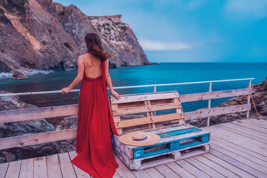 Side view a Young beautiful sensual woman in a red long dress posing on a rock high above the sea during sunrise. Girl on the nature on blue sky background. Fashion photo.