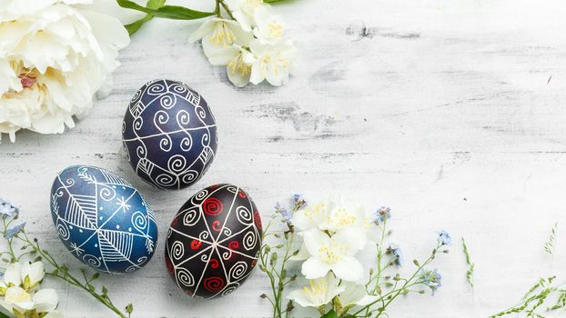 Three handmade painted Easter eggs with spring flowers. Ukrainian pysanka decorated with wax-resist dyeing technique on white shabby wooden background with empty space for text