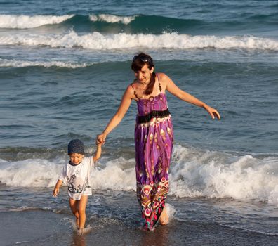 A young woman in a long summer dress walks with her little son among the waves on the seashore