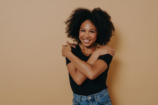 Image of beautiful pleased young dark skinned female with voluminous afro hairstyle, embracing herself with beaming smile, isolated over studio beige background with copy space. Self love concept