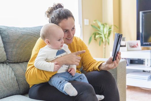 young woman with her baby making a video call with her mobile phone in her living room. Family time.