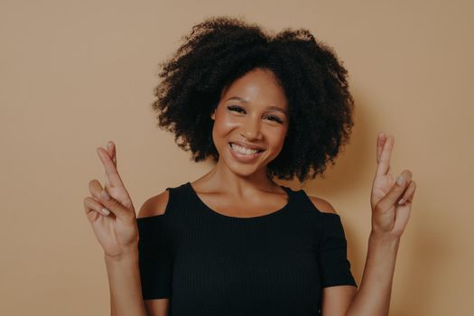 Portrait of happy young african woman standing isolated over dark beige studio background with copy space, holding fingers crossed for good luck broadly smiling. Positive emotions and body language