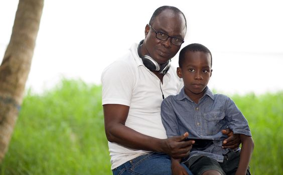 concept of family, paternity and people - father and son sitting in the garden and using a mobile phone