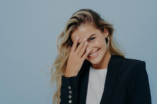 Happy shy young blonde woman in formal clothes covering her face with hand, at loss for words because of joyful emotions, does not know what to say, standing isolated on blue background