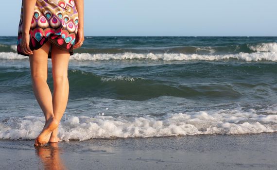 Young beautiful woman in a summer dress stands in the sea water on the sandy shore and enjoys the waves and the sea breeze