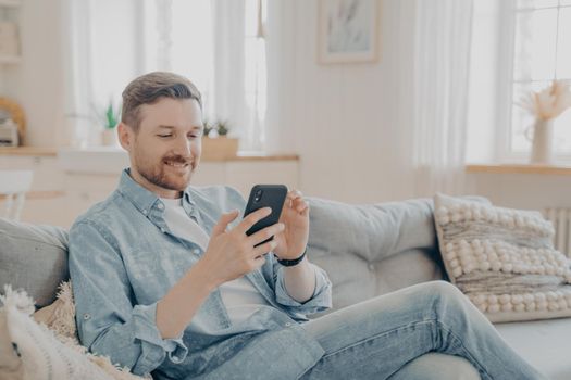 Happy smiling bearded man using smartphone device while sitting on sofa in living room, staying on couch and reading messages on mobile phone while relaxing during leisure time at home