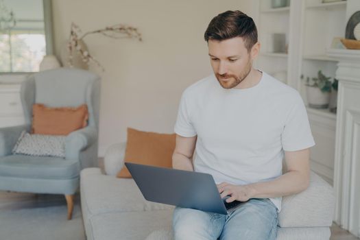 Serious young man using laptop computer for remote work from home, focused freelancer guy reading incoming emails or chatting online, researching or studying while sitting on couch in living room