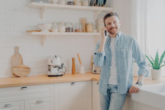 Handsome smiling young bearded man wearing casual clothes talking on mobile phone in kitchen while working online remotely from home, having successful conversation with client about signing contract