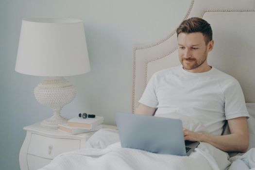 Young handsome cheerful smiling man freelancer in casual pajama outfit sitting in bed in morning and working remotely on laptop at home while resting in cozy bed. Freelance concept