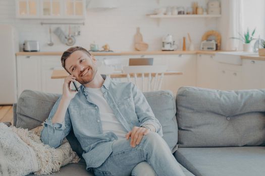 Smiling guy sits on couch at home having fun while talking on modern cellphone gadget, happy young male relaxing on sofa in living room while making conversation with his girlfriend