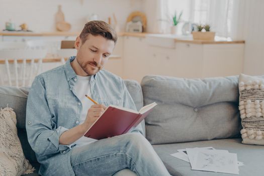 Busy young man dressed in casual clothes writing in red notebook with pencil while sitting on comfortable couch with crossed legs, looking focused, kitchen in blurred background