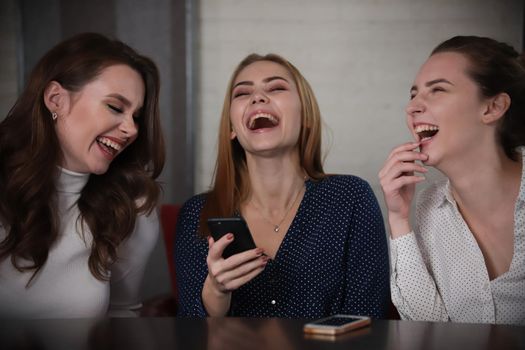 Three young beautiful women sitting in a cafe looking on telephone, laughing. Portrait