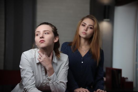 Portrait of two beautiful young women sitting at the table looking at camera over lights in background. Portrait