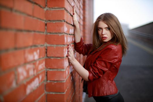 Portrait of young woman in black leggings and red leather jacket standing near red brick wall