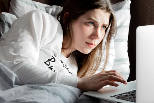 Young cute girl on a sunny day in a white jacket lies on the bed and looks into the laptop.