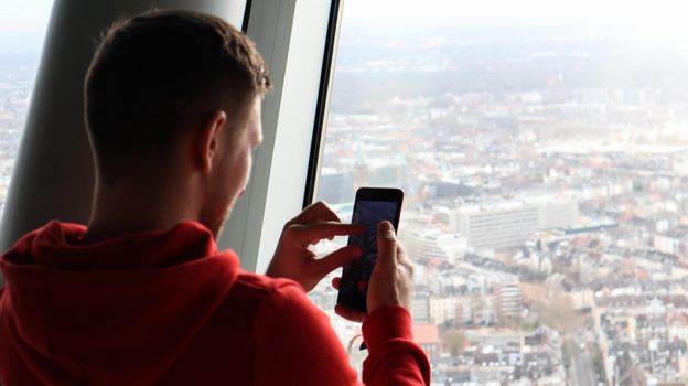 A young man makes a photo of the cityscape on a smartphone. Tourism concept. A man photographs a city from an observation deck through a glass from a height