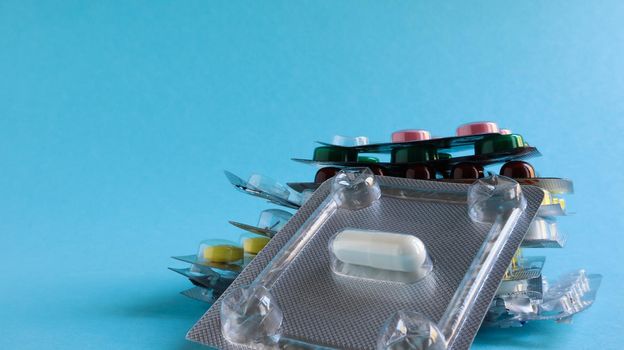 Packs of pills of various shapes and colors are stacked on a blue background. Capsules are packaged in blister packs. Different medicines. Drug treatment. Health photo. Copy space