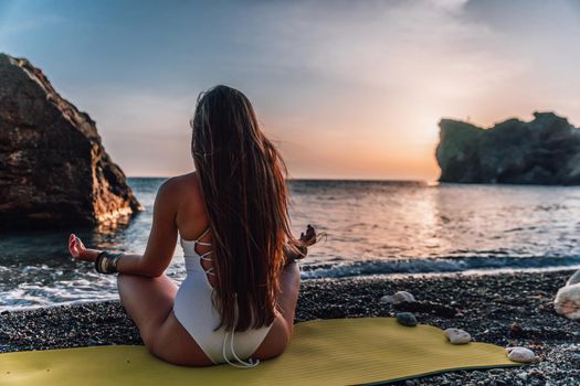 Young woman in swimsuit with long hair practicing stretching outdoors on yoga mat by the sea on a sunny day. Women's yoga fitness pilates routine. Healthy lifestyle, harmony and meditation concept.