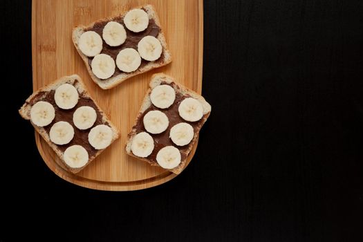 Three banana white bread toasts spread with chocolate butter that lie on a chopping board on a dark background. top view with area for text