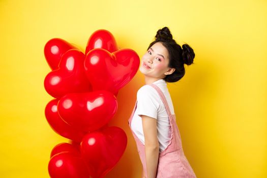 Valentines day and relationship concept. Fashionable asian woman posing near red hearts balloons, surprise for lover on special date, yellow background.