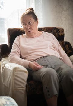 Pastimes. An elderly caucasian woman in glasses enjoys her hobby sitting in a armchair and knitting on the background of the window. Portrait