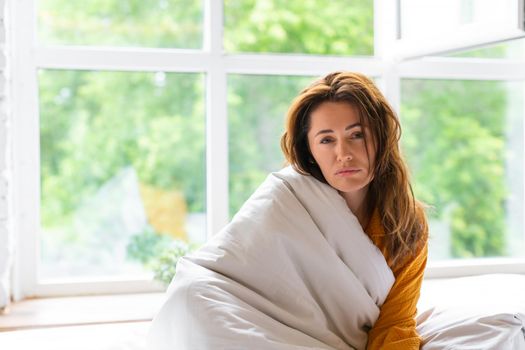 40 year old woman is sick. She sits on the bed covered with a blanket and sadly looks at the camera.