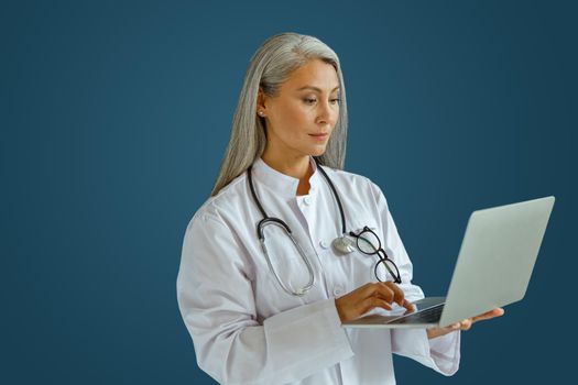 Concentrated grey haired woman doctor with stethoscope works on laptop standing on blue background in studio. Telemedicine patient service