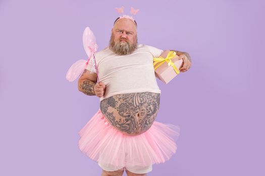 Funny bearded plump male person in fairy costume with magic stick and gift box grimaces on purple background in studio