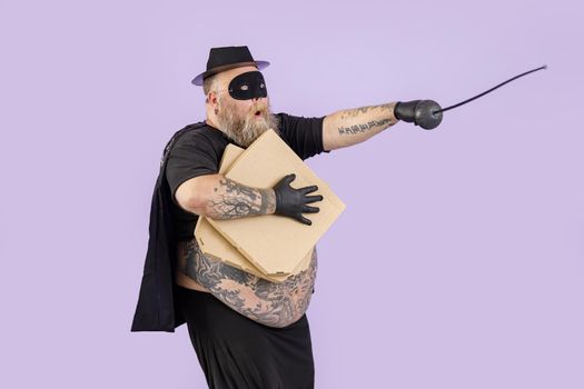Emotional mature fat man in Zorro costume with epee holds cardboard boxes of pizza standing in bellicose pose on purple background in studio