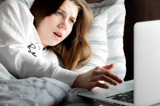 A young cute girl on a sunny day in a white jacket lying on the bed and looking at the laptop in surprise.