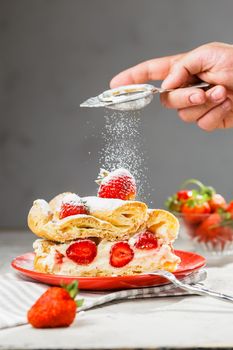 A piece of strawberry cake on a red plate. Man's hand with sieve icing sugar powder from above. Grey background, vertical composition