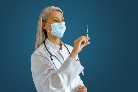 Professional Asian woman therapist in white robe with disposable mask holds syringe with medicine standing on blue background in studio. Vaccination day