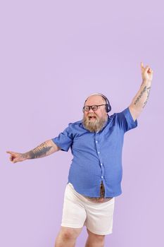 Positive mature bearded man with overweight in tight shirt and shorts enjoys favourite music with headphones dancing on purple background in studio
