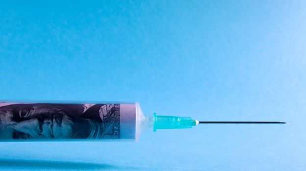 A roll of 100 dollars money inserted into a white medical hypodermic needle on a blue background. The concept of health care costs, credit, investment. Copy space for your text or logo