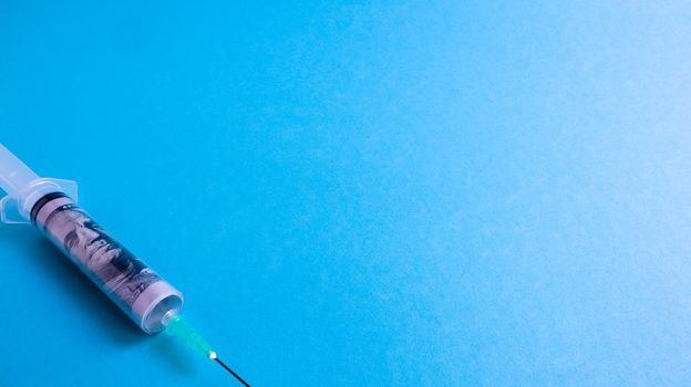A roll of 100 dollars money inserted into a white medical hypodermic needle on a blue background. The concept of health care costs, credit, investment. Copy space for your text or logo
