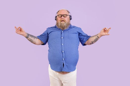 Tranquil mature bearded obese man in tight shirt meditates while listens to relaxing music with headphones on purple background in studio