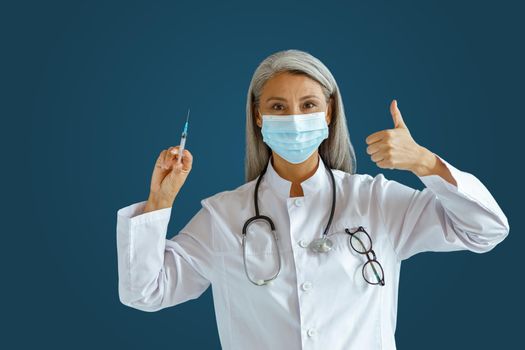 Mature Asian woman doctor in white coat with medical mask holds syringe and shows thumb up standing on blue background in studio. Vaccination day