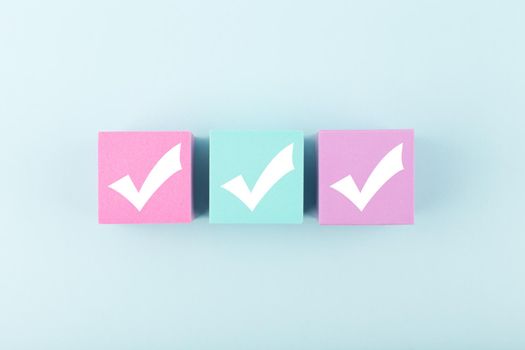Three checkmarks on multicolored cubes in a row in the middle of bright pastel blue background. Concept of questionary, checklist, to do list, planning, business or verification. Modern minimal composition