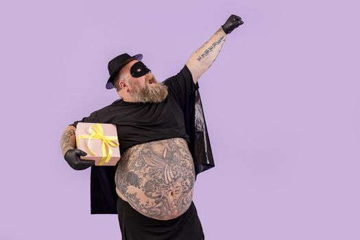 Funny emotional gentleman with overweight wearing carnval hero costume with cape and mask holds present on purple background in studio