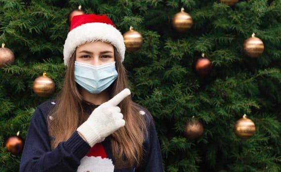 Close up Portrait of woman wearing a santa claus hat and medical mask with emotion and show the finger. Against the background of a Christmas tree. Coronavirus pandemic