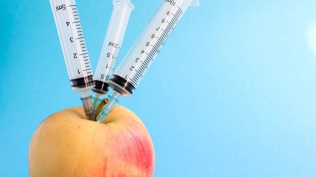 Three different medical syringes sticking deep into an apple on a blue background. The fruit is strewn with syringes. A ripe red apple pierced from all sides by syringes. Healthcare or cosmetology