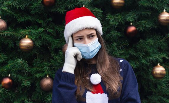 Choosing a gift. Close up Portrait of woman wearing a santa claus hat and medical mask with emotion. Against the background of a Christmas tree. Coronavirus pandemic