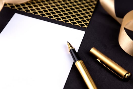 Gold pen, ribbon, paper clips and stationery on a black background with a white sheet of paper.