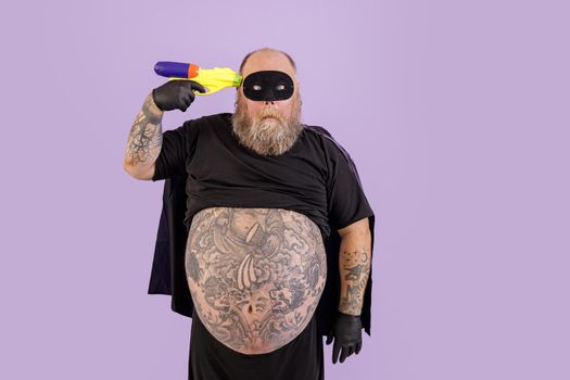 Funny obese man wearing hero costume with cape and mask holds toy blaster to temple posing on purple background in studio