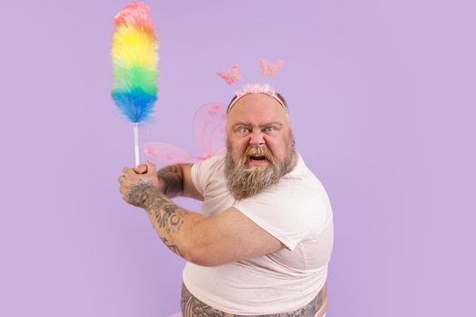 Funny angry bearded plump man in fairy suit with wings attacks with fluffy dust brush on purple background in studio