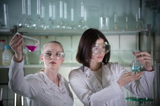 Chemical laboratory. Two young woman in work glasses holding different flasks with liquids in it. Looking at the flask. Portrait