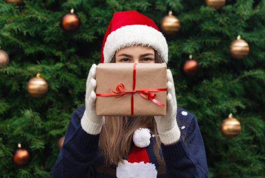 A Christmas gift. A young girl wearing a santa claus hat gives a gift made of craft paper with a red ribbon. Against the background of a Christmas tree
