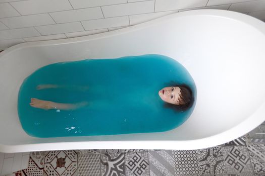 Baby boy swims on his back in blue water in bathtub. Top view of adorable little kid lying relaxing during hygiene routine